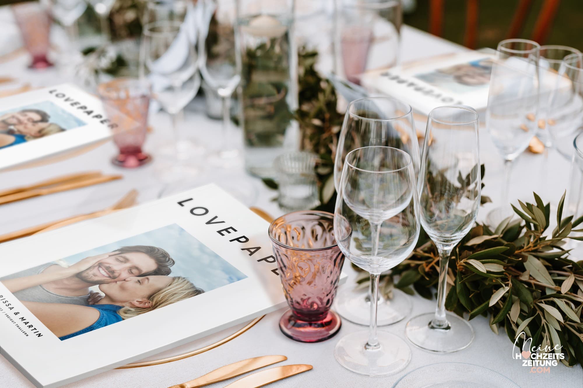 Gedeckter Hochzeitstisch mit Gläsern, Olivenzweigen und goldenem Besteck, an jedem Platz liegt ein Magazin „Lovepaper“ mit Paarfoto.