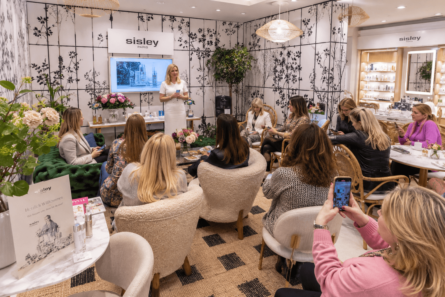 Referentin stellt im Sisley-Paris-Raum einer kleinen Runde sitzender Gäste Produkte vor, eine Teilnehmerin fotografiert mit dem Handy