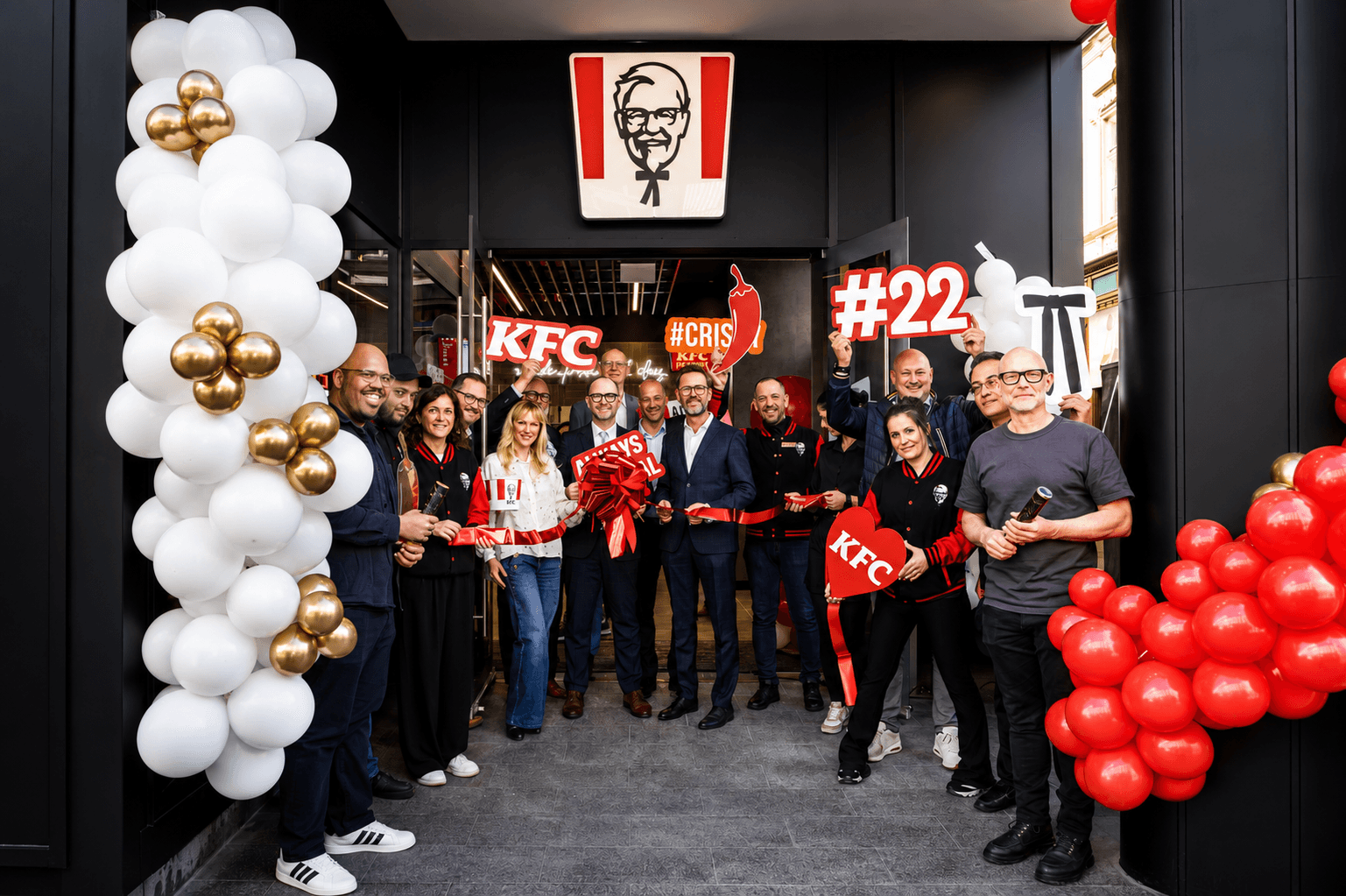 Eröffnung einer KFC-Filiale: Eine Gruppe steht mit rotem Band vor dem Eingang, flankiert von weiß-goldenen und roten Ballons.