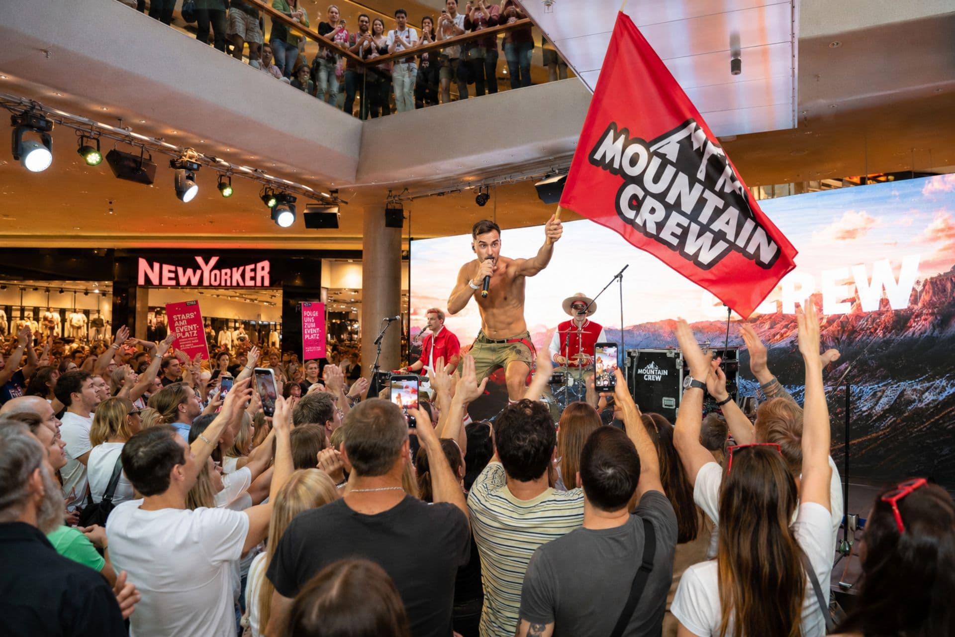 Ein Sänger schwenkt auf einer Bühne im Einkaufszentrum eine rote „Mountain Crew“-Fahne, davor jubelt das Publikum, einige mit Handys.
