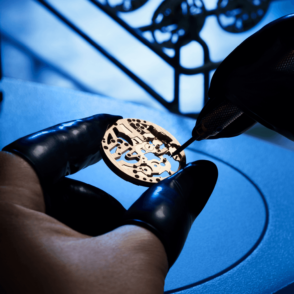 Eine Hand mit schwarzen Fingerlingen hält ein durchbrochenes, goldfarbenes Uhrwerkteil, eine zweite führt ein feines Werkzeug – im blauen Licht.