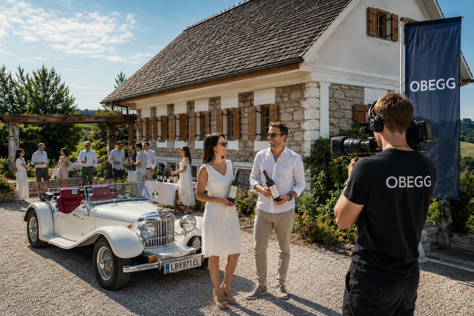Kameramann im OBEGG-Shirt filmt einen Mann und eine Frau mit Weinflaschen vor einem Steinhaus und einem weißen Oldtimer.