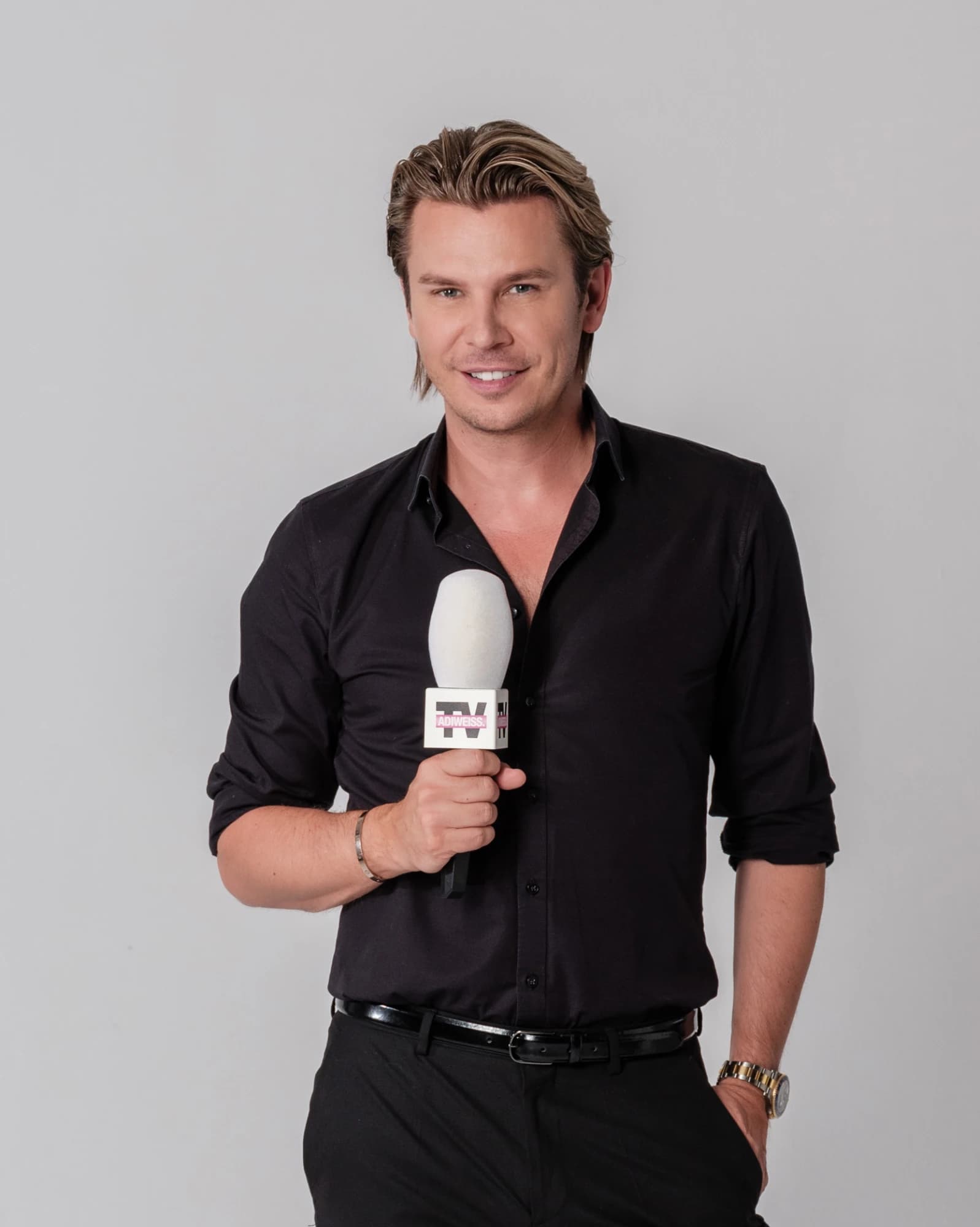 Adi Weiss, Moderator von ADIWEISS.TV, lächelt im schwarzen Hemd und hält ein Mikrofon mit TV-Logo vor grauem Hintergrund