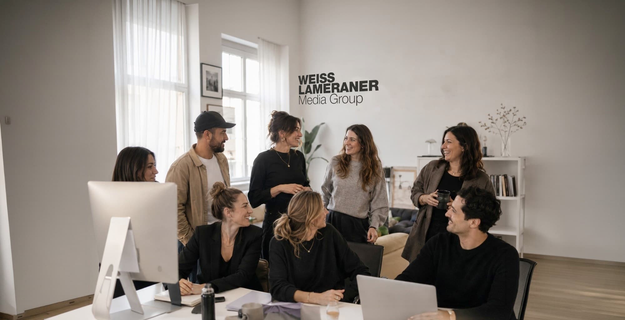 Acht Teammitglieder unterhalten sich lachend in einem hellen Büro, an der Wand das Logo Weiss & Lameraner Media Group.