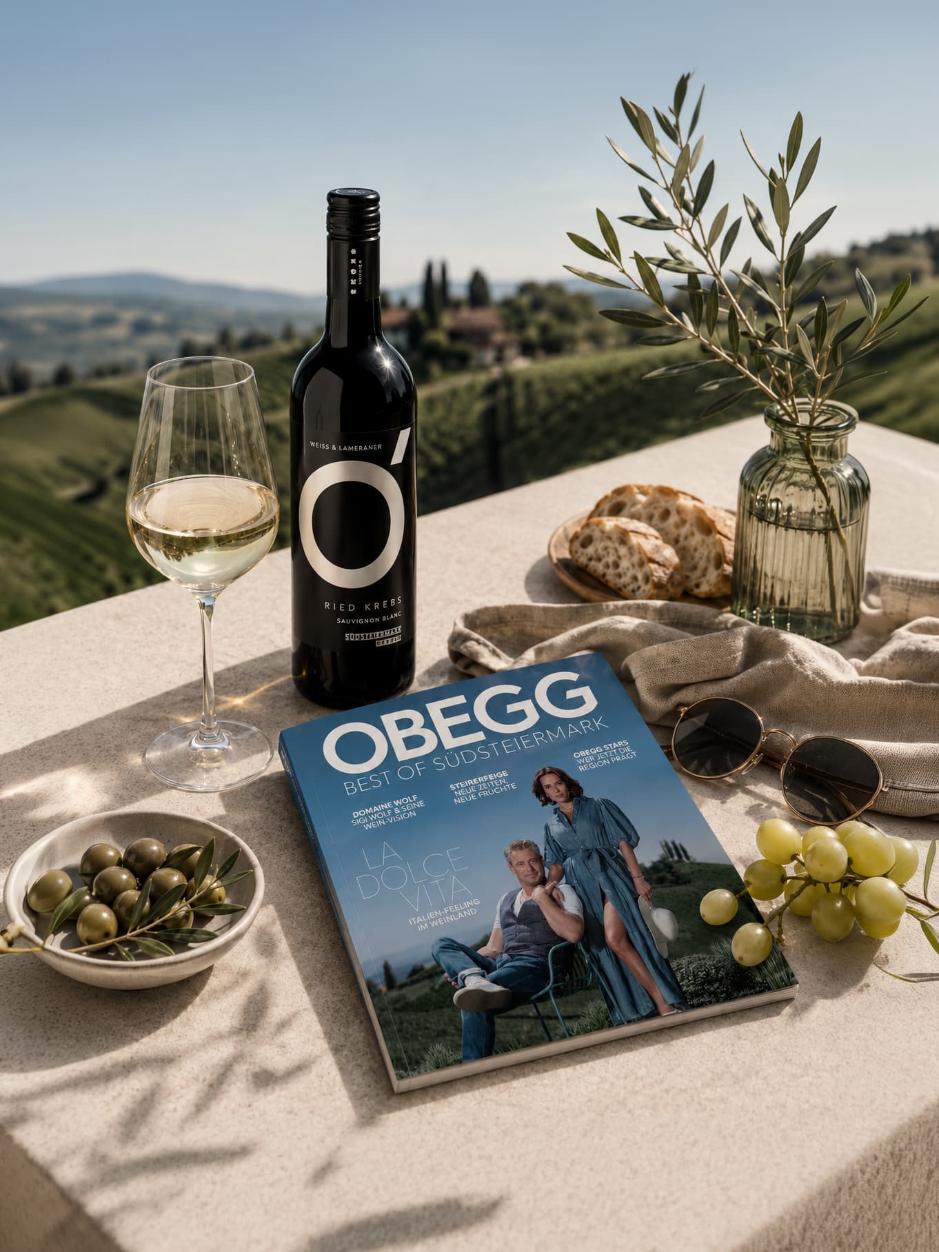 OBEGG-Magazin „Best of Südsteiermark“ auf einem Steintisch mit Weißwein, Oliven und Trauben vor einer Weinberglandschaft